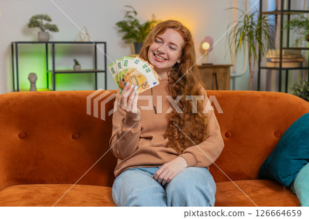 Successful rich young Caucasian woman holding euro bill money fan sitting on sofa couch at home Successful rich young Caucasian woman holding euro bill money fan sitting on sofa couch at home 126664659