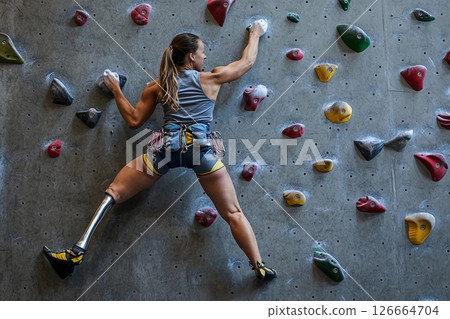 Female athlete with prosthetic leg climbs indoor rock wall in training session at climbing gym 126664704