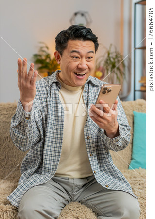 Asian middle-aged man at home celebrating online game victory, shocked by good news on phone 126664785