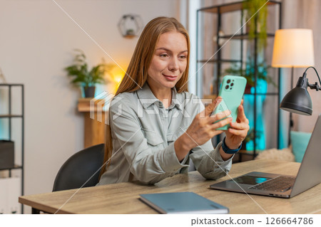 Happy female freelancer sitting at desk using smartphone texting smiling in living room at home 126664786