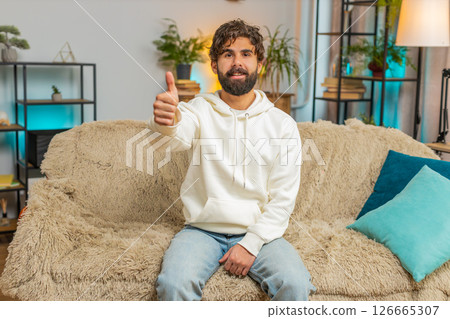 Happy excited Indian young man sitting on sofa couch looking at camera and showing thumbs up sign 126665307