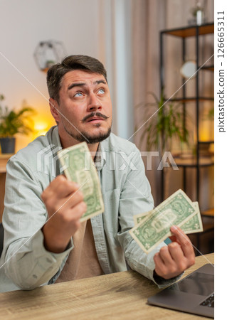 Upset Caucasian man freelancer counting dollar cash insufficient amount of money sitting at table Upset Caucasian man freelancer counting dollar cash insufficient amount of money sitting at table 126665311