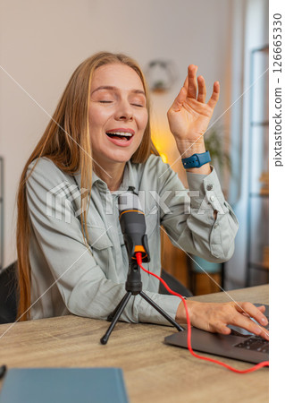 Mature positive Caucasian woman blogger singer using laptop while singing into microphone at table Mature positive Caucasian woman blogger singer using laptop while singing into microphone at table 126665330