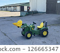Green toy tractor with yellow scoop parked on a concrete surface in front of a barn, showcasing a playful design for children and outdoor activities 126665347