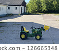 Green toy tractor parked on a concrete surface near a building, surrounded by trees, showcasing a playful outdoor environment with ample copy space for design 126665348