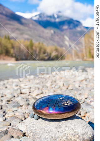 Steel tongue drum resting on a stone by the river in the Altai mountains. Concept of elemental harmony, raw sound energy, and nature as a sacred space for music and silence Steel tongue drum resting on a stone by the river in the Altai mountains. Concept of elemental harmony, raw sound energy, and nature as a sacred space for music and silence 126668445