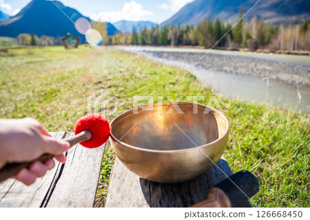 Point of view of hand playing a Tibetan singing bowl near mountain river in Altai. Concept of personal ritual, sound healing, and spiritual harmony with wild nature. 126668450