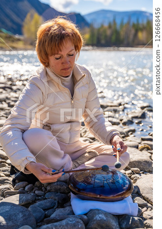Woman playing a steel tongue drum near a mountain river in Altai, surrounded by wild nature. Concept of spiritual retreat, sound healing, and ancient connection to earth Woman playing a steel tongue drum near a mountain river in Altai, surrounded by wild nature. Concept of spiritual retreat, sound healing, and ancient connection to earth 126668456