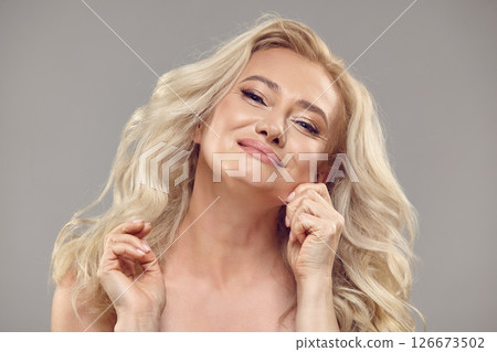 Attractive senior woman pulling cheeks and smiling looking at camera. Close up beauty portrait of happy mature lady with hands on cheek over isolated grey background. Attractive senior woman pulling cheeks and smiling looking at camera. Close up beauty portrait of happy mature lady with hands on cheek over isolated grey background. 126673502