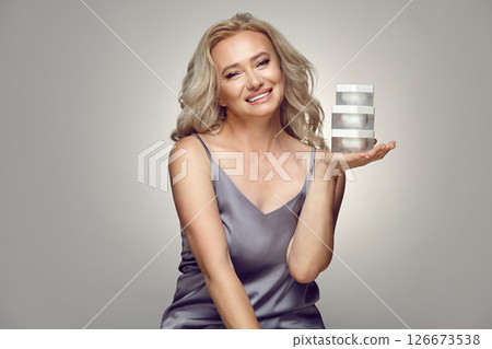 Caucasian mature woman holding jars of moisturizer near her face on a grey isolated. 126673538
