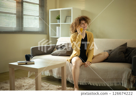 Curly haired woman in a mustard cardigan enjoys a cheerful phone call in living room 126674222