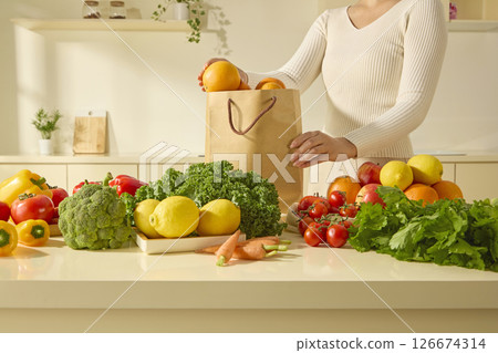 A person holds a recyclable paper bag of produce in a bright kitchen filled with vegetables such as kale, bell peppers and cherry tomatoes on the counter. Fresh vegetables on beige table in kitchen A person holds a recyclable paper bag of produce in a bright kitchen filled with vegetables such as kale, bell peppers and cherry tomatoes on the counter. Fresh vegetables on beige table in kitchen 126674314