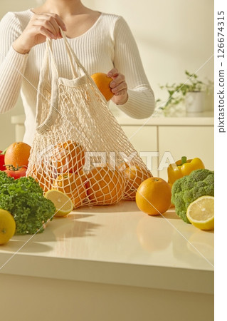 A woman places a reusable bag of fresh fruits and vegetables on a kitchen island, surrounded by colorful produce such as apples, celery, cauliflower, tomatoes and lemons. Kitchen background 126674315