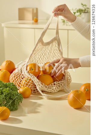 Close-up of hands putting oranges into a mesh bag in a warm, natural-light kitchen, highlighting eco-friendly grocery shopping and healthy food choices 126674316
