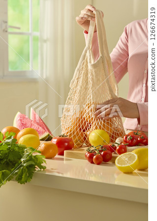 A person holds a reusable bag filled with bright oranges in a sunlit kitchen, emphasizing sustainability and fresh vitamin-rich fruit.Fresh vegetables in the kitchen, promotional photo A person holds a reusable bag filled with bright oranges in a sunlit kitchen, emphasizing sustainability and fresh vitamin-rich fruit.Fresh vegetables in the kitchen, promotional photo 126674319