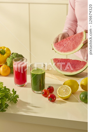Woman pouring watermelon juice in a glass next to a variety of fresh greens, lemon slices, and cherry tomatoes in a bright home kitchen. Colorful fresh vegetables in the kitchen, healthy food concept 126674320