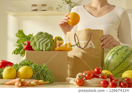 A woman picks up an orange from a paper grocery bag, surrounded by colorful and fresh produce. Modern kitchen, healthy organic food A woman picks up an orange from a paper grocery bag, surrounded by colorful and fresh produce. Modern kitchen, healthy organic food 126674324