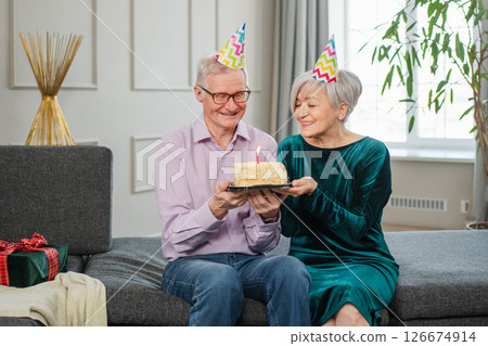 Make a wish. Family senior couple in party hat cap celebrating birthday anniversary together at home. Old woman blowing out burning candles on birthday cake. Old man wishes wife happy birthday 126674914