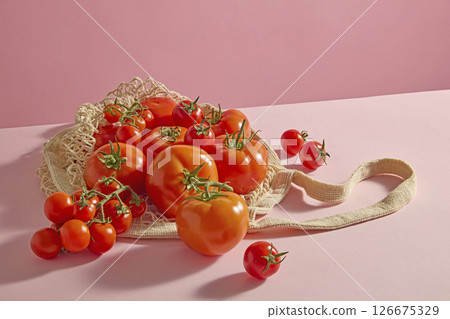 A side-view image of fresh vegetables, including cherry tomatoes, spilling out of a mesh bag onto a pastel pink surface. Gentle shadows and highlights create a soft, natural feel with ample copy space 126675329