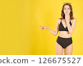 Shocked bikini model with hand on mouth and finger pointing left, standing on bright yellow background, concept image for marketing, sale announcements, and lifestyle promotion templates 126675527