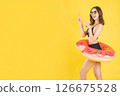 Young woman in bikini with donut float and sunglasses pointing up against vibrant yellow studio background, smiling with cheerful expression, perfect for summer ads, banners, and promotions 126675528