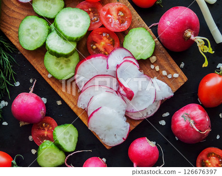 Red cherry tomatoes, radish, herbs and cucumbers sprinkled with sea salt on the wooden board on black table, top view of vegetables with water drops. 126676397
