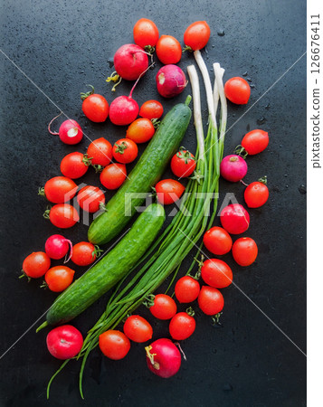 Fresh red cherry tomatoes, green leek, radish and cucumber on the black table background, top view of vegetables covered with water drops. Fresh red cherry tomatoes, green leek, radish and cucumber on the black table background, top view of vegetables covered with water drops. 126676411
