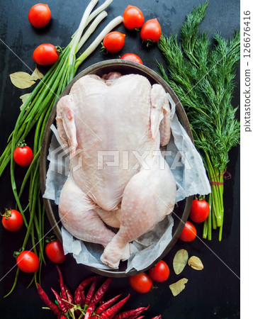Whole raw chicken laying in the pan with parchment on the kitchen table preparing for baking, surrounded by fresh vegetables and herbs, top view. 126676416