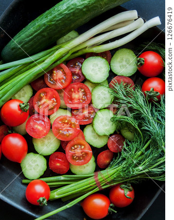 Fresh red cherry tomatoes, green leek, radish and cucumber in pan on the black background, top view of vegetables covered with water drops. Fresh red cherry tomatoes, green leek, radish and cucumber in pan on the black background, top view of vegetables covered with water drops. 126676419