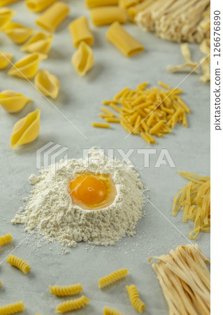 Variety of uncooked pasta with flour and egg yolk forming a circle on a gray background Variety of uncooked pasta with flour and egg yolk forming a circle on a gray background 126676890