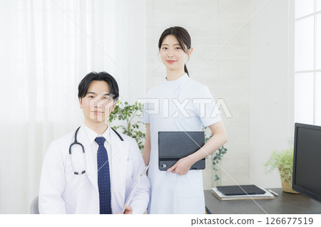 A man in a white coat talking to a nurse in an examination room 126677519