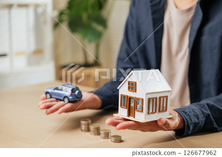 Person displays car and house models above coin stacks, illustrating money-saving strategies for auto and real estate investments. Perfect for business, banking, and personal financial goals. 126677962