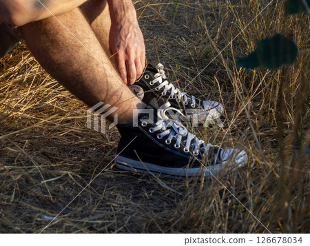 Individual sitting in dry grass wearing black high top sneakers while enjoying a peaceful moment in nature during late afternoon sunlight 126678034