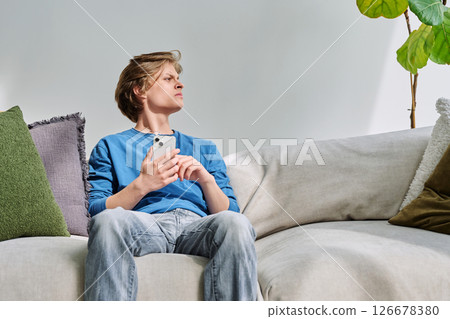Handsome serious teenage guy with smartphone sitting on couch at home Handsome serious teenage guy with smartphone sitting on couch at home 126678380