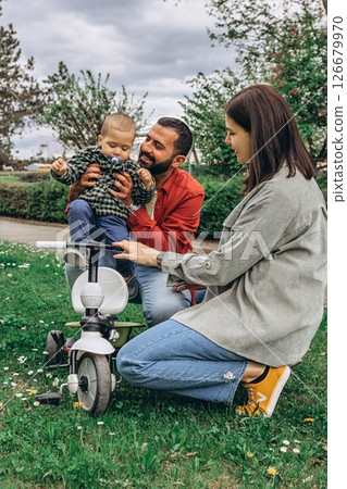 A mom and dad with their 2-year-old son in the park. The toddler is riding a small bike, enjoying family time outdoors filled with joy and learning 126679970