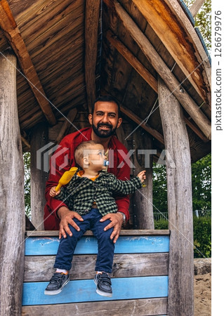 Father and young child playing together in a wooden playhouse at an outdoor playground. The child pours sand from a yellow scoop, surrounded by natural wood textures and greenery Father and young child playing together in a wooden playhouse at an outdoor playground. The child pours sand from a yellow scoop, surrounded by natural wood textures and greenery 126679976