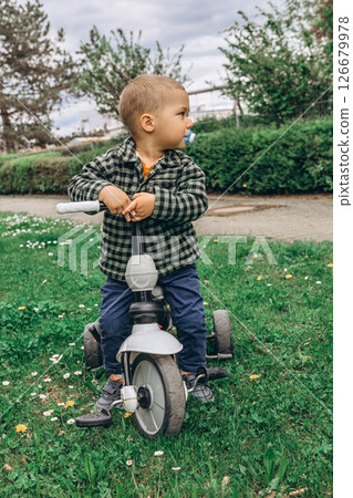 A 2-year-old boy riding a small bike in the park with a pacifier in his mouth. A sweet and candid moment of early childhood, outdoor activity, and toddler development in a natural setting A 2-year-old boy riding a small bike in the park with a pacifier in his mouth. A sweet and candid moment of early childhood, outdoor activity, and toddler development in a natural setting 126679978