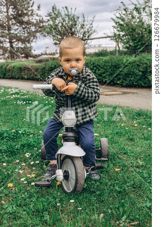 A 2-year-old boy riding a small bike in the park with a pacifier in his mouth. A sweet and candid moment of early childhood, outdoor activity, and toddler development in a natural setting A 2-year-old boy riding a small bike in the park with a pacifier in his mouth. A sweet and candid moment of early childhood, outdoor activity, and toddler development in a natural setting 126679984