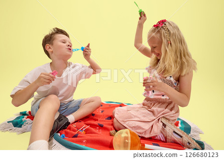 Close-up of children, boy in white t-shirt, girl in pink dress with red bow, playing with soap bubbles, laughing with joy, sitting on colorful towel Close-up of children, boy in white t-shirt, girl in pink dress with red bow, playing with soap bubbles, laughing with joy, sitting on colorful towel 126680123