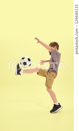 Boy lifting leg to balance ball in classic soccer move, wearing shorts and sneakers, yellow background enhances focus on movement and coordination Boy lifting leg to balance ball in classic soccer move, wearing shorts and sneakers, yellow background enhances focus on movement and coordination 126680135