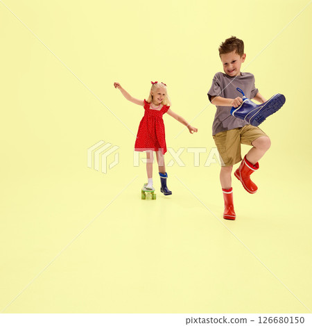 Girl and boy having fun putting on mismatched rubber boots, laughing and walking playfully with quirky props against yellow background Girl and boy having fun putting on mismatched rubber boots, laughing and walking playfully with quirky props against yellow background 126680150