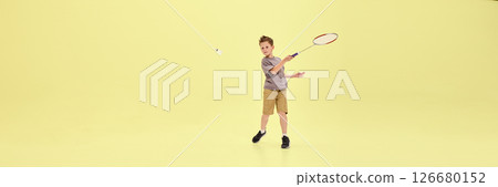 Boy lunges forward swinging racket at flying shuttlecock, fully immersed in game against pastel yellow background with copy space. Banner 126680152
