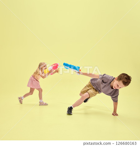 Girl and boy energetically chasing each other with water guns, joyful game captured mid-movement against yellow background Girl and boy energetically chasing each other with water guns, joyful game captured mid-movement against yellow background 126680163