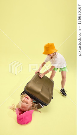 Boy playfully trying to close suitcase over laughing girl lying inside against pastel backdrop, bright colors and expressive faces create comic joyful moment Boy playfully trying to close suitcase over laughing girl lying inside against pastel backdrop, bright colors and expressive faces create comic joyful moment 126680167