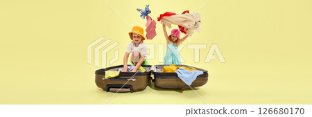 Two kids cheerfully tossing clothes from open suitcase, expressing excitement and anticipation of summer holidays on yellow background. Banner Two kids cheerfully tossing clothes from open suitcase, expressing excitement and anticipation of summer holidays on yellow background. Banner 126680170