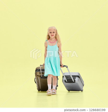 Girl in turquoise dress walks forward pulling two rolling suitcases, looking straight with confident smile against clean yellow studio background. Girl in turquoise dress walks forward pulling two rolling suitcases, looking straight with confident smile against clean yellow studio background. 126680199