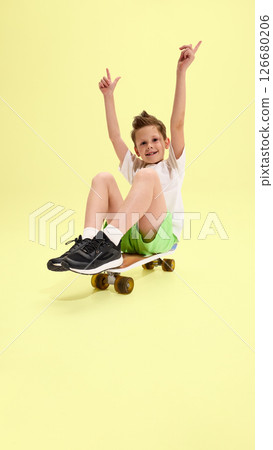 Child sitting on skateboard with raised arms, smiling widely and showing excitement, dressed in green shorts and white tee against yellow background 126680206