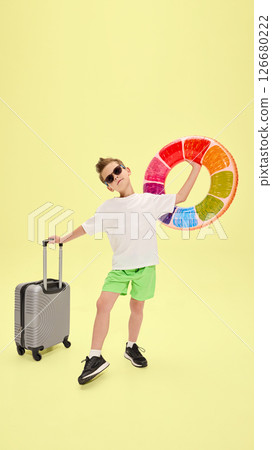 Cool boy in stylish pose holding suitcase and colorful inflatable ring, standing on one leg and acting playful against bright yellow studio background 126680222
