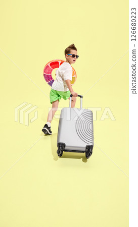 Boy walking forward and looking back while pulling suitcase, holding inflatable swim ring, ready for beach adventure against yellow background 126680223