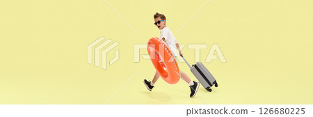 Playful boy in sunglasses walking with suitcase and swim ring, preparing for summer journey with happy and carefree attitude against yellow background Playful boy in sunglasses walking with suitcase and swim ring, preparing for summer journey with happy and carefree attitude against yellow background 126680225